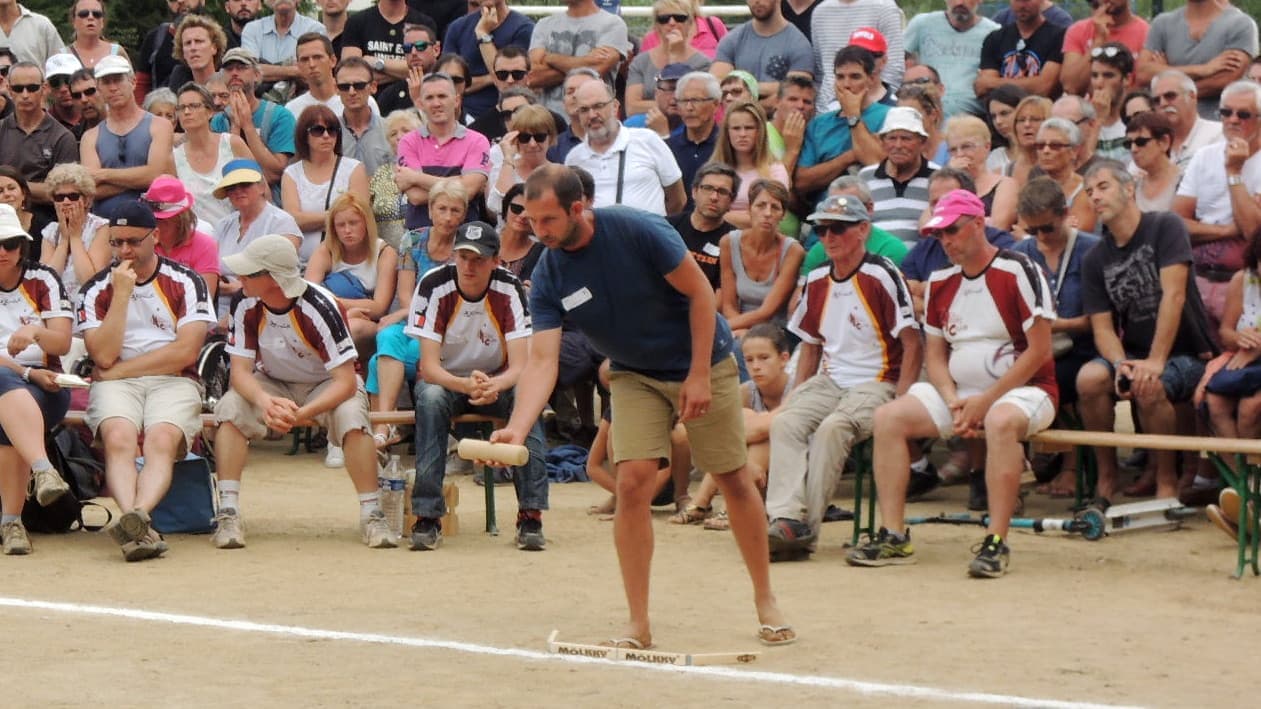 A player about to throw underarm, both feet behind a wooden marker laid on the ground, with seated teammates and a crowd behind.